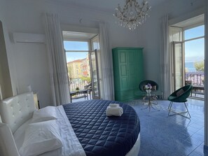 Une chambre avec un grand lit, une armoire verte, un balcon donnant sur des immeubles et un lustre.