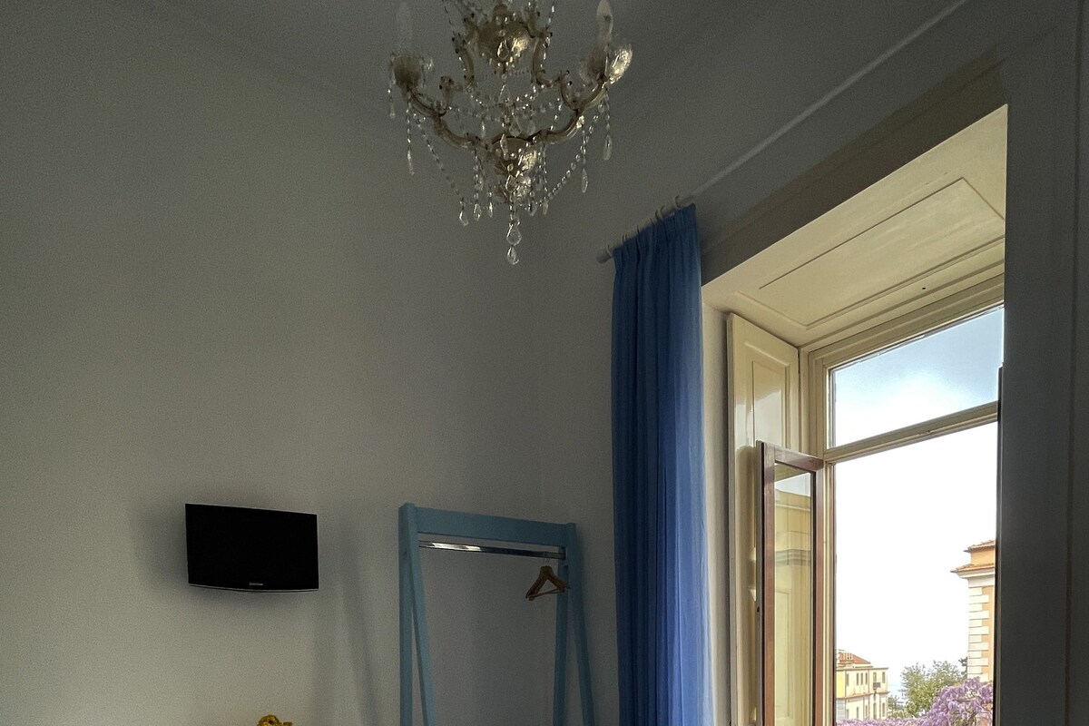 Ein Hotelzimmer mit einem Bett, einem Schreibtisch mit Stuhl, einem Fernseher, einem Fenster mit Blick auf Gebäude und einer Kristallleuchtre.