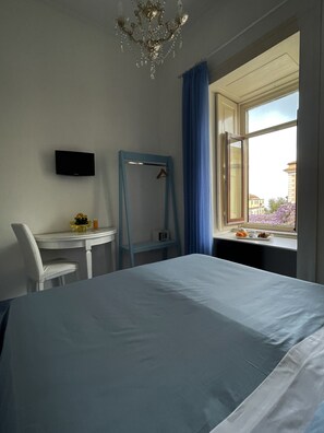 Ein Hotelzimmer mit einem Bett, einem Schreibtisch mit Stuhl, einem Fernseher, einem Fenster mit Blick auf Gebäude und einer Kristallleuchtre.