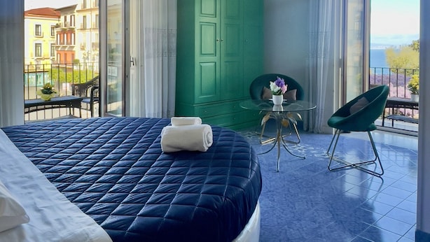 A bedroom with a large bed, a green wardrobe, a balcony with a view of buildings, and a chandelier. - Ambasciata Sorrento Relais (Sorrento)