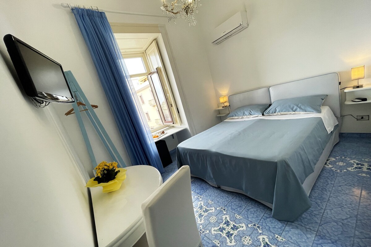 Ein Hotelzimmer mit Bett, Schreibtisch, Stuhl, Spiegel, einem Fenster mit Vorhängen und einer Chandelier.