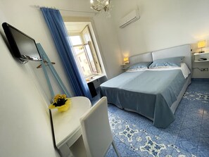 Ein Hotelzimmer mit Bett, Schreibtisch, Stuhl, Spiegel, einem Fenster mit Vorhängen und einer Chandelier.