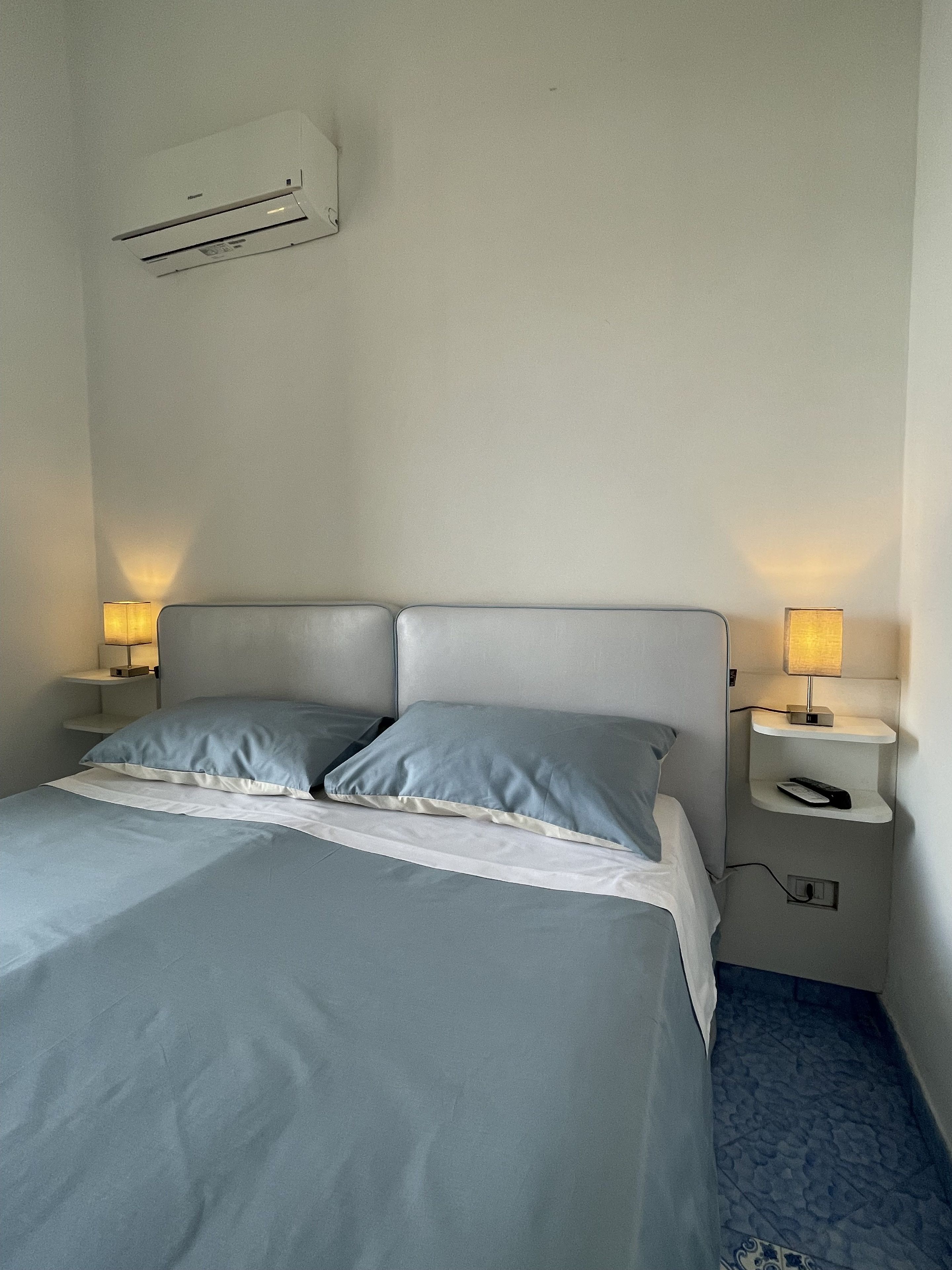 A neatly made bed with two pillows, a bedside table with a lamp, and a wall-mounted air conditioner.