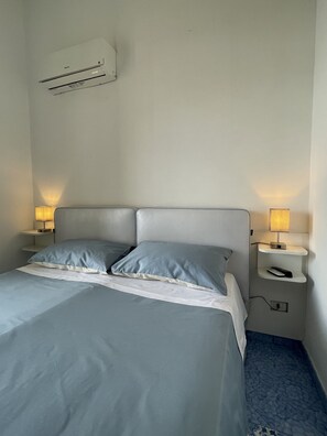 Ein ordentlich bezogenes Bett mit zwei Kissen, ein Nachttisch mit Lampe und eine Wand-Klimaanlage.