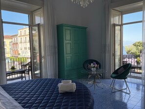 Ein Schlafzimmer mit einem großen Bett, einem grünen Kleiderschrank, einem kleinen Tisch mit einer Vase, einem Stuhl und einem Balkon mit Blick auf Gebäude und einen Baum.