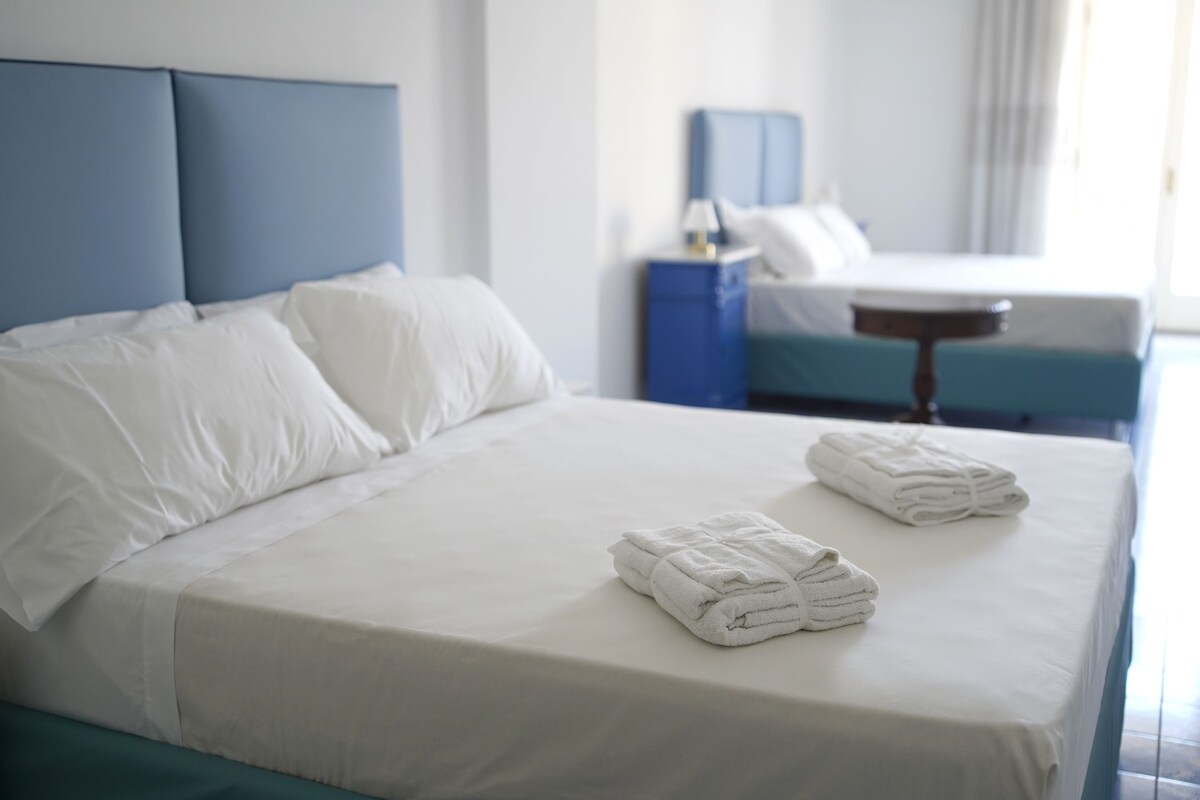 Un lit bien fait, avec du linge de lit blanc et deux serviettes pliées, une tête de lit bleue et une table de chevet avec une lampe.