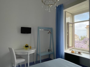 Une chambre d’hôtel avec un lit, un bureau et une chaise, un téléviseur fixé au mur, une fenêtre donnant sur des bâtiments et des fleurs, et une chaise bleue avec un porte-manteau.