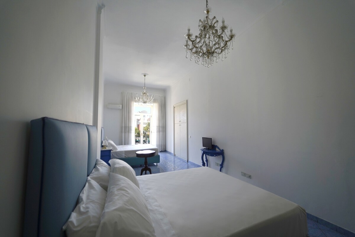 Ein Hotelzimmer mit einem großen Bett, einem kleinen runden Tisch, einem Schreibtisch mit Computer, einer Kristallleuchter und einem Fenster mit Vorhängen.