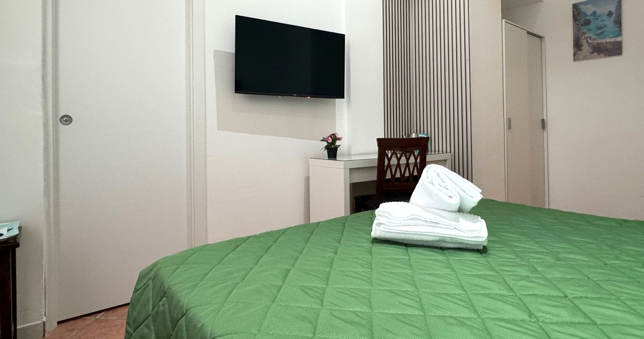 Habitación de hotel con edredón verde, televisor de pantalla plana en la pared, una mesita con planta en maceta y una silla.