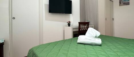 Habitación de hotel con edredón verde, televisor de pantalla plana en la pared, una mesita con planta en maceta y una silla.