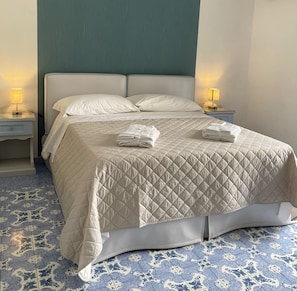 Ein ordentlich gemachtes Bett mit einer gesteppten Bettdecke, zwei Kissen und gefalteten Handtüchern auf der Seite. Zwei Nachttische mit Lampen stehen bereit, und der Boden ist mit gemusterten Fliesen ausgelegt.