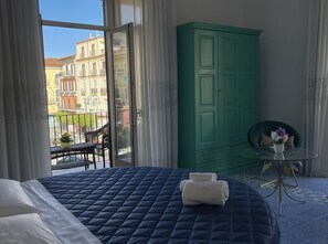 Quarto com cama, guarda-roupa verde, mesa redonda pequena com um vaso, e vista de edifícios por uma grande janela.