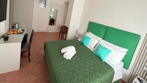 Hotelkamer met een groen bed, witte kussens, een klein bureau met waterkoker, sinaasappels en een potplant.
