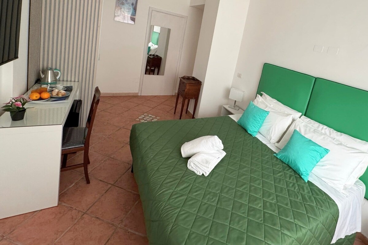 Ein Hotelzimmer mit einem grünen Bett, weißen Kissen, einem kleinen Schreibtisch mit Wasserkocher, Orangen und einer Topfpflanze.