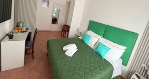 Ein Hotelzimmer mit einem grünen Bett, weißen Kissen, einem kleinen Schreibtisch mit Wasserkocher, Orangen und einer Topfpflanze.