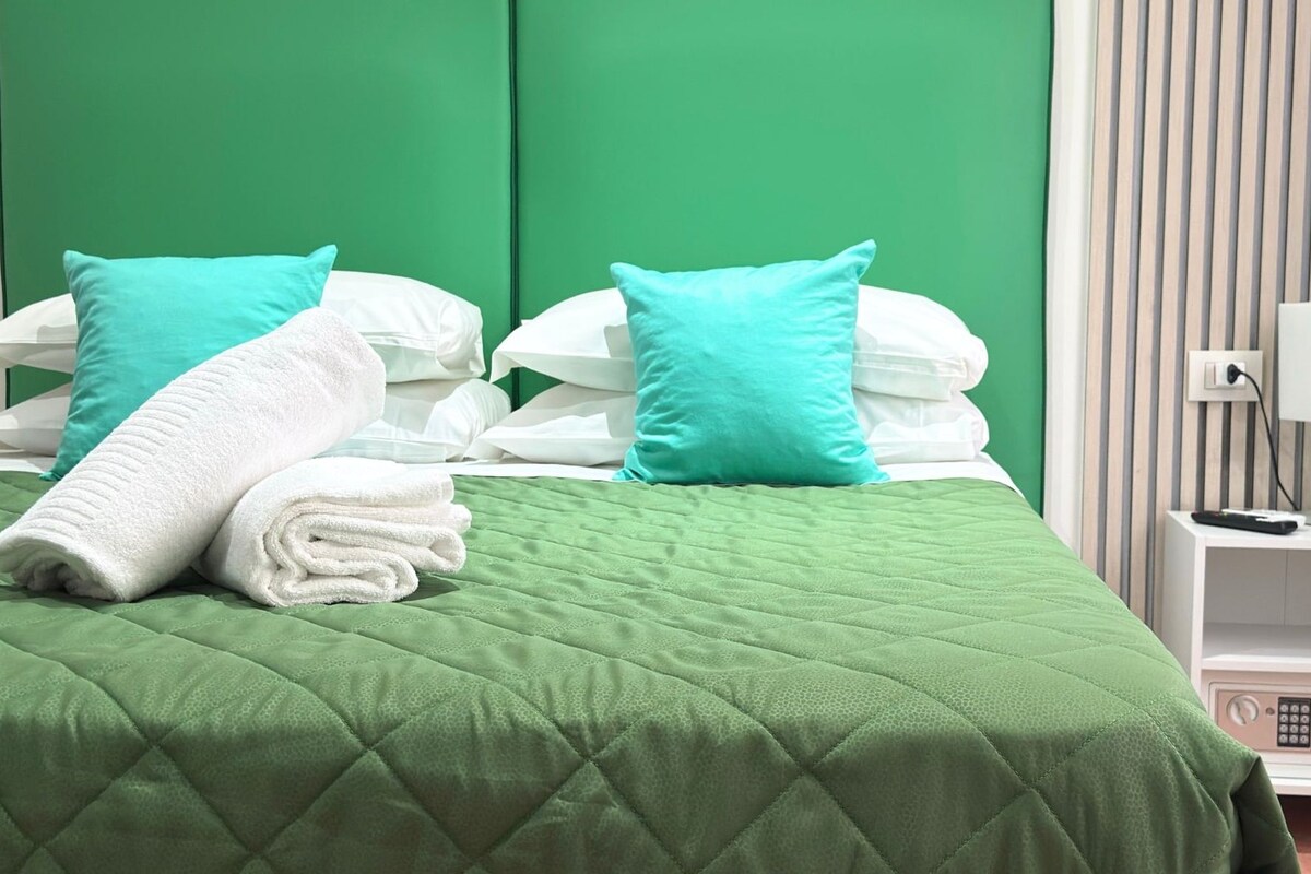 Un lit bien fait, avec une literie verte, des oreillers blancs et une serviette blanche pliée.