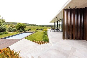 Une terrasse extérieure moderne, dotée d’une piscine, d’une terrasse en bois et d’un espace salon offrant une vue sur la campagne.