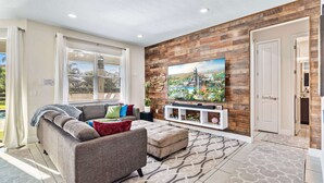 A modern living room with a sectional sofa, a flat-screen TV mounted on a wooden wall, and a large window with blinds.