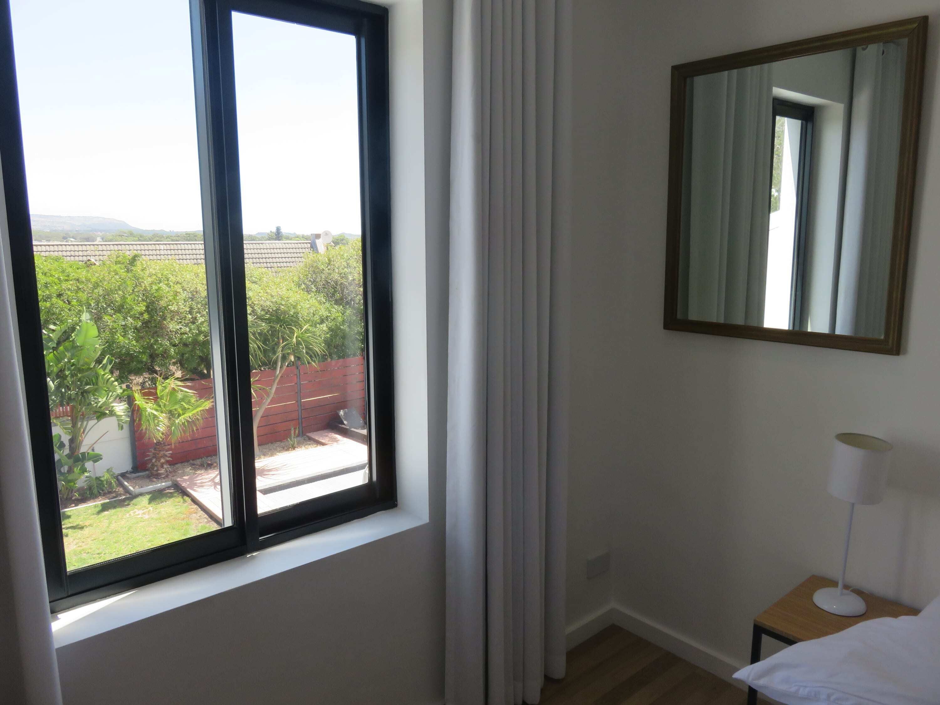 Une chambre avec une grande fenêtre, un miroir, une table de chevet avec une lampe, et une vue sur un jardin et des bâtiments.