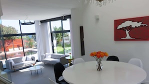 A modern living room with a white sofa, a dining table with chairs, and a vase of flowers. There is a large window with curtains, a chandelier, and a painting on the wall. - Cape white villa - Family & Friends suite (Cape Town)