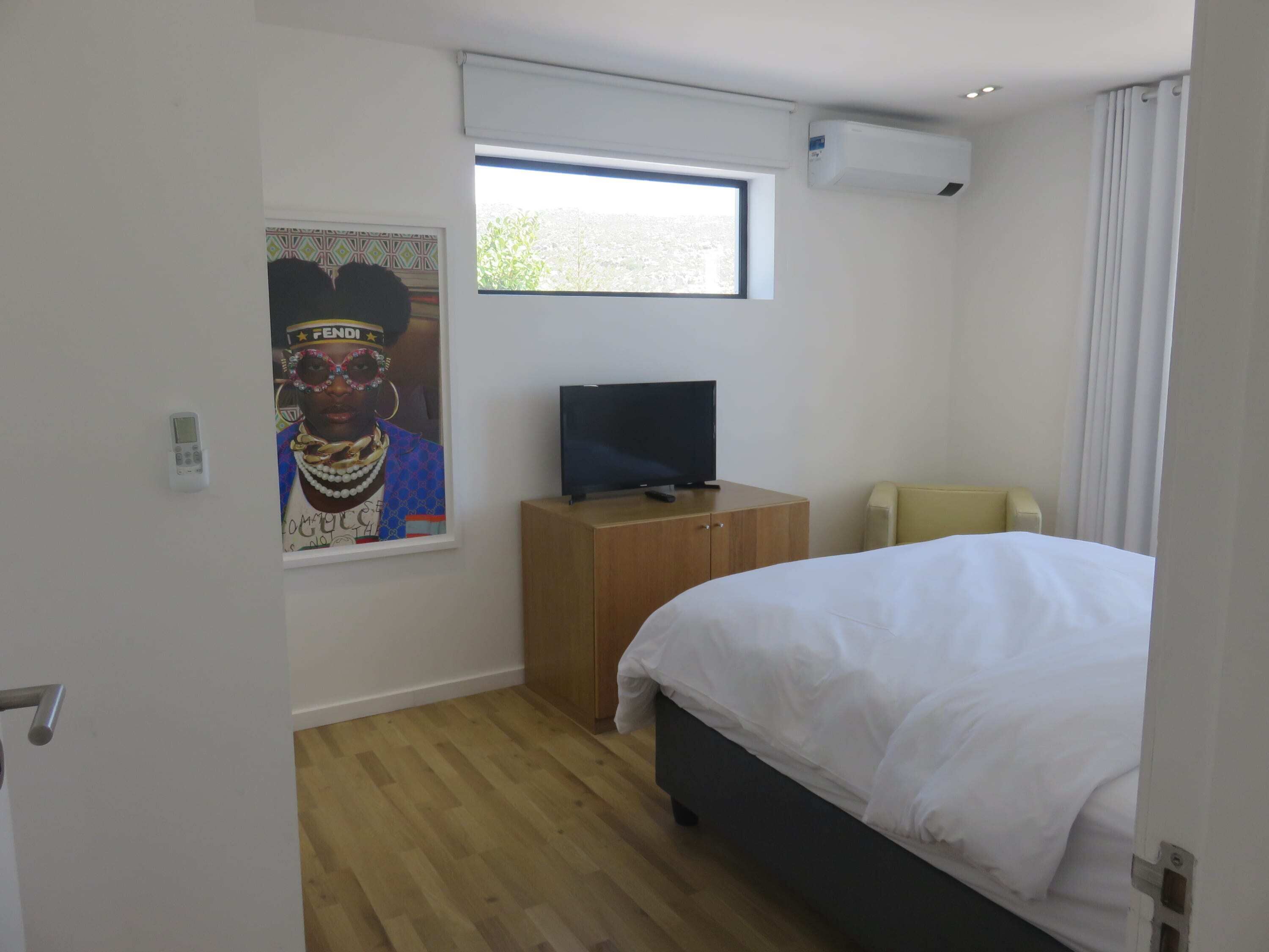 A bedroom with a bed, a TV on a wooden cabinet, a window with a view of trees, and an air conditioning unit.