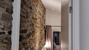 Un dormitorio con pared de piedra, una cama con ropa de cama blanca y vistas a un paisaje urbano a través de una ventana.