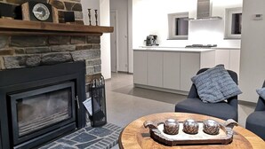 Una sala moderna con chimenea, una mesa de centro redonda de madera con objetos decorativos, un sofá y una cocina al fondo.