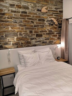 A bedroom with a stone wall, a bed with white bedding, a nightstand with a clock, and a wooden nightstand.