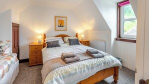 A bedroom with a sloped ceiling, a bed with striped bedding, two bedside tables with lamps, a framed picture on the wall, and a window with a view of greenery. - Carraig Dhubh North - UK47125 (Whiting Bay)