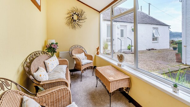 A cozy room with wicker furniture, a decorative wreath, and a view of a white house and greenery outside. - Carraig Dhubh North - UK47125 (Whiting Bay)