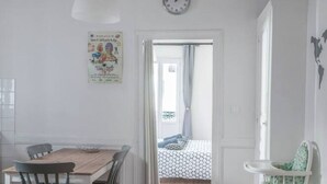 Une pièce comprenant une table à manger en bois, des chaises, un luminaire blanc au plafond, une horloge murale et un lit recouvert d’une couvre-lit à motifs.