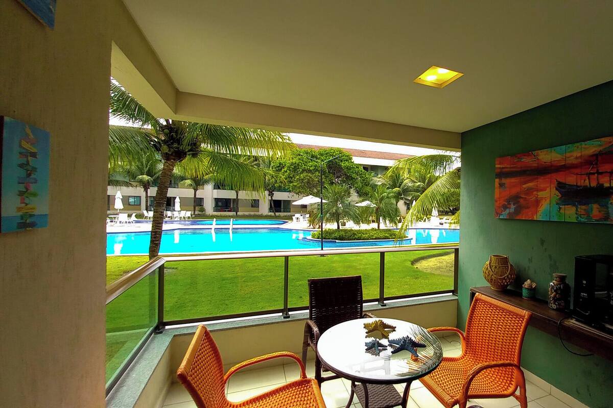 A balcony with a view of a swimming pool, wicker chairs, a small table, and a painting on the wall.