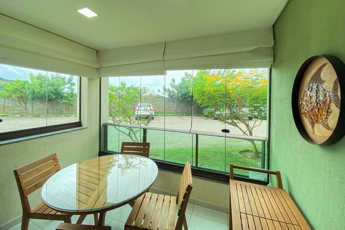 A room with a round glass table and wooden chairs, large windows with a view of a parking area and trees, and a wall-mounted decorative piece.