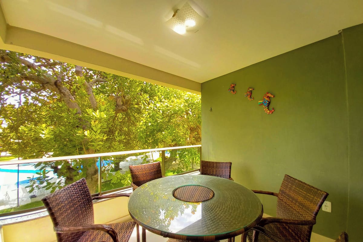 A balcony with wicker furniture, a glass table, and a view of trees and a pool.