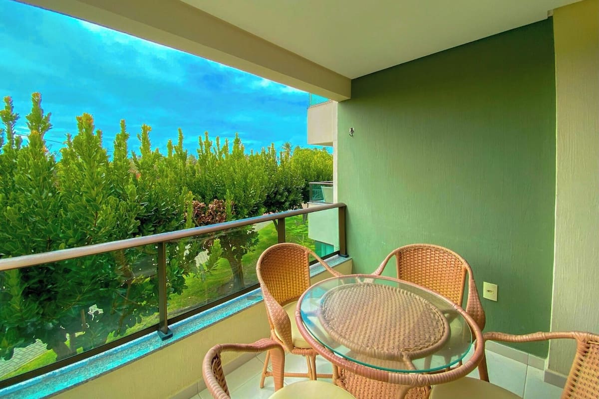 A balcony with wicker chairs and a glass table, overlooking a garden with green foliage and a clear sky.