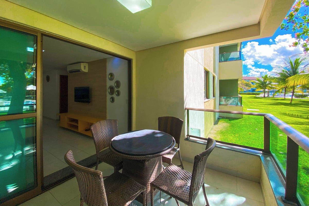 A balcony with wicker furniture, a flat-screen TV, and a view of a well-maintained lawn and buildings.