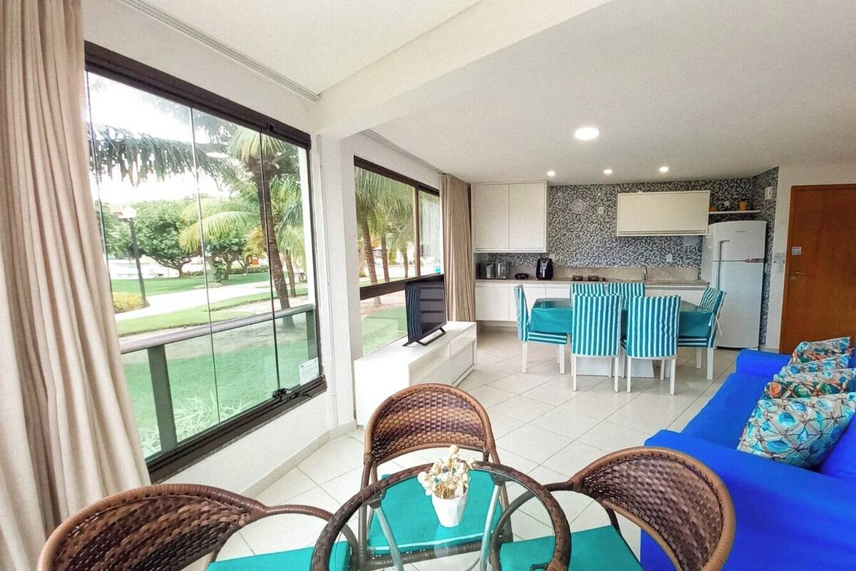 A modern living room with a blue sofa, wicker chairs, and a dining table with chairs. A kitchen with a tiled backsplash and a refrigerator is visible in the background.