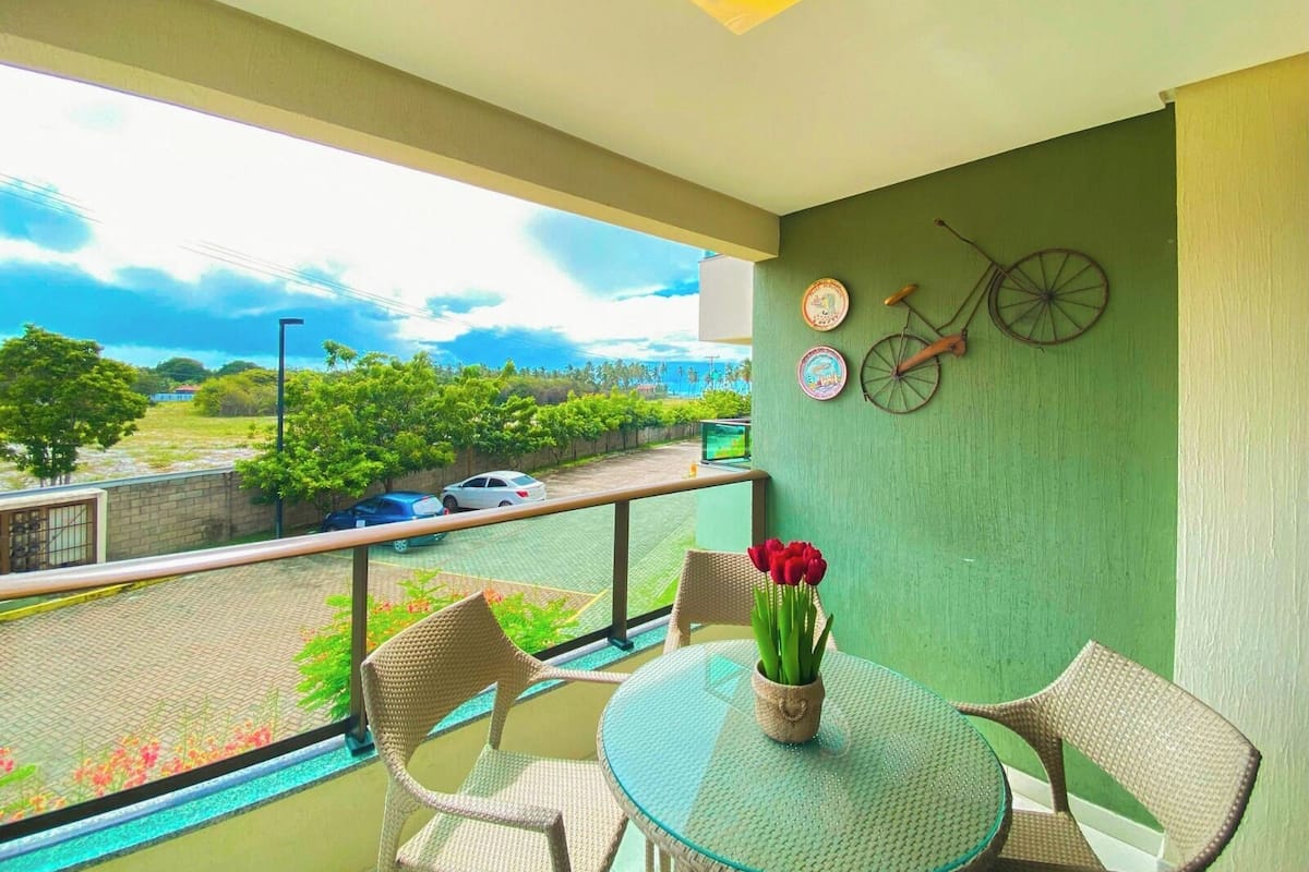 A balcony with wicker chairs, a glass table, and a vase of red flowers. There are two decorative plates and a bicycle on the wall.