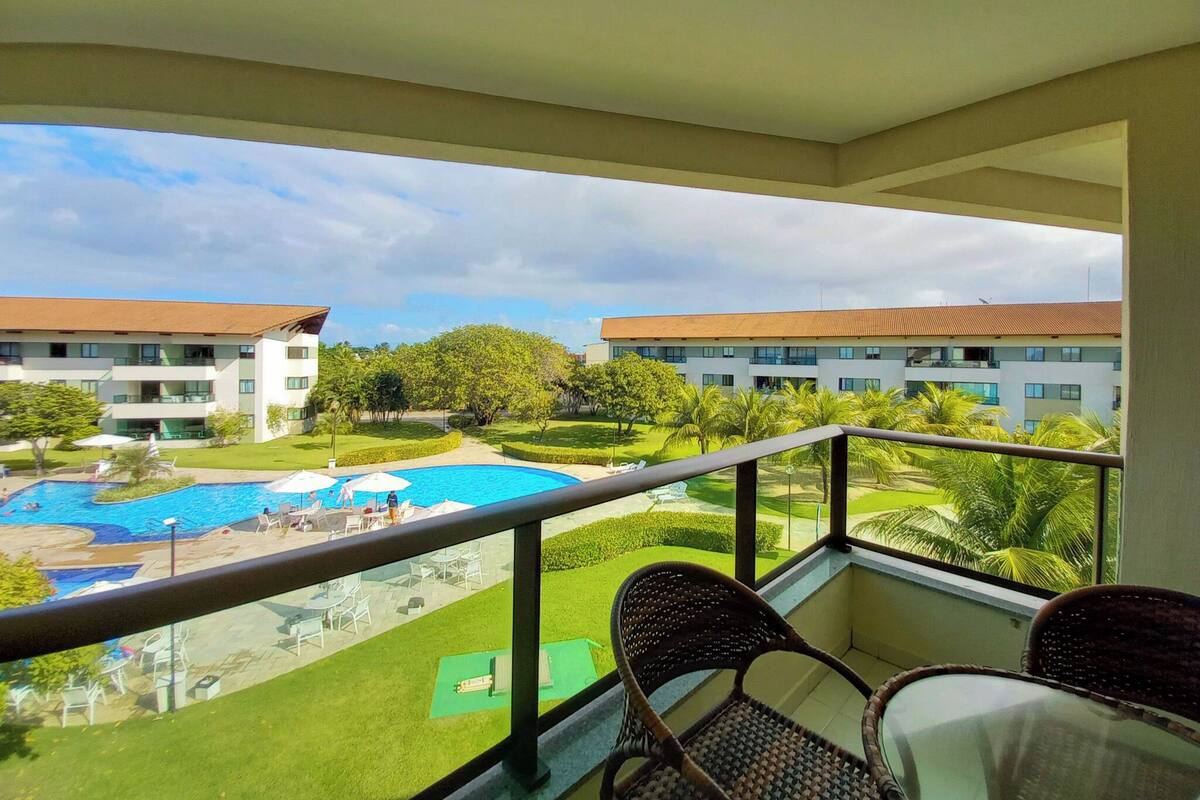 A balcony with wicker chairs, a pool, and a view of a building with a red roof.