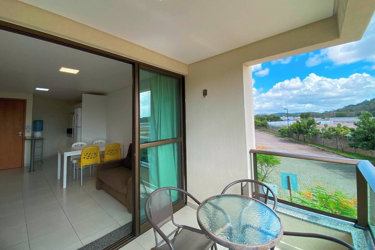 A balcony with a glass table and chairs, a sliding glass door leading to an interior room, and a view of a green landscape with trees and a clear sky.