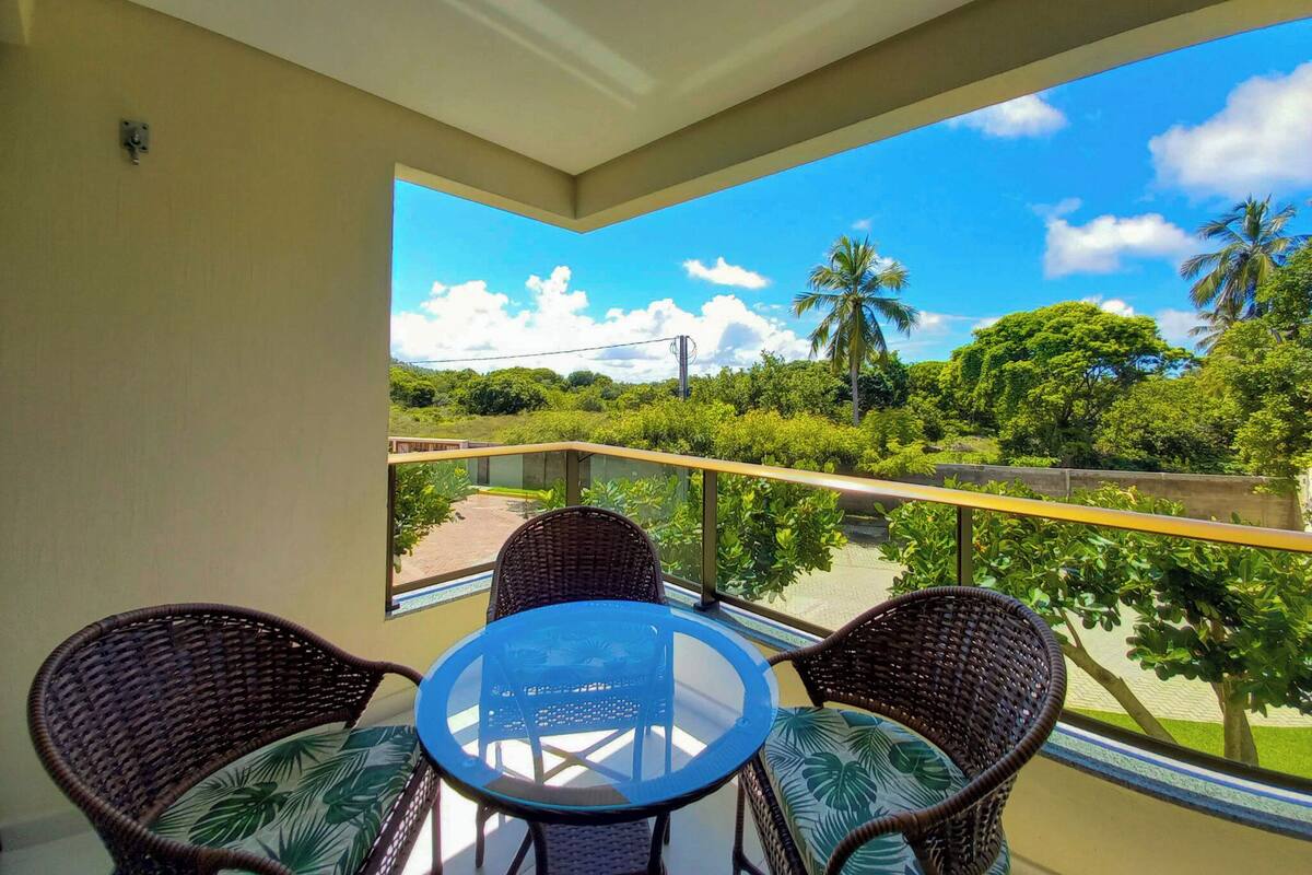 A balcony with wicker furniture and a glass table, overlooking a lush landscape with palm trees and a clear sky.