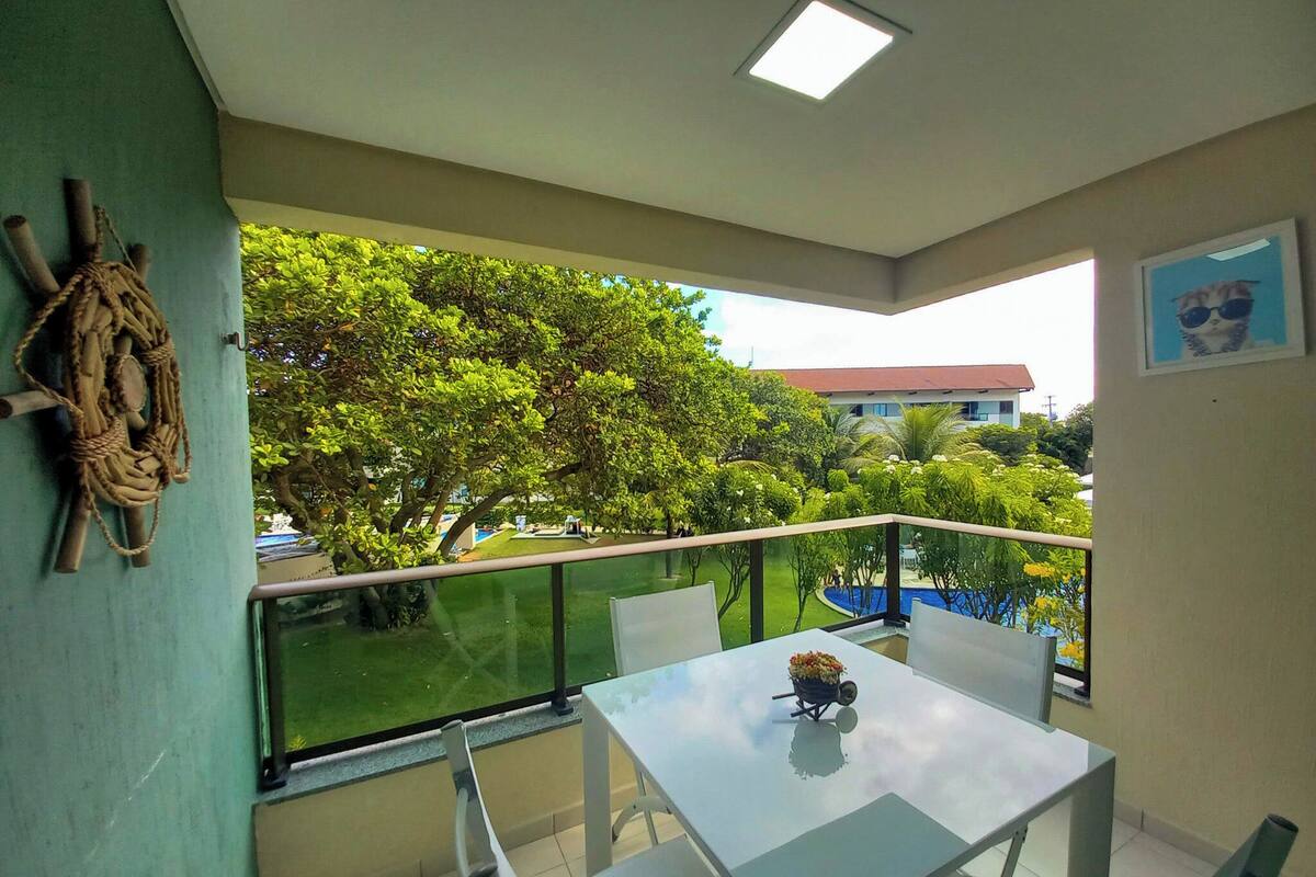 A balcony with a glass railing, a table with a flower pot, and a view of lush greenery and buildings.