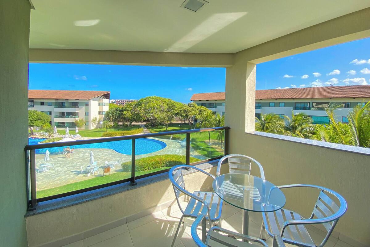 A balcony with a glass table and chairs, overlooking a swimming pool and a building with a red roof.