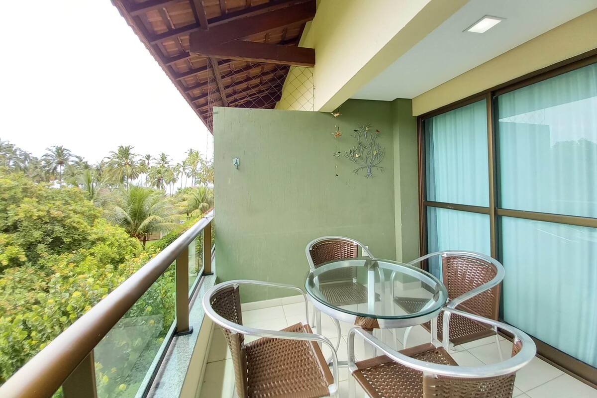 A balcony with wicker chairs and a glass table, overlooking a lush green landscape.