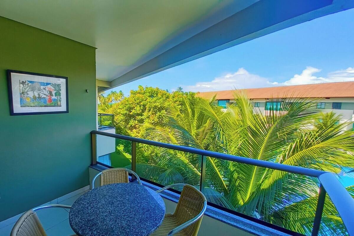 A balcony with a table and chairs, a framed picture on the wall, and a view of palm trees and buildings.