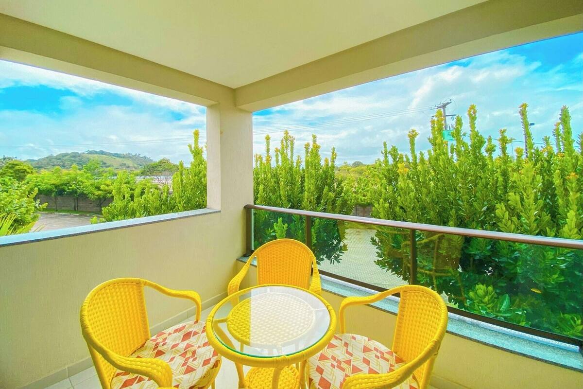 A balcony with yellow wicker chairs and a small glass table, overlooking a lush green landscape with tall trees and a clear sky.