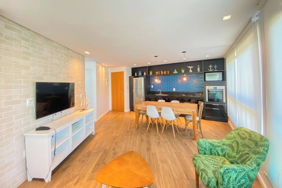 A modern living room with a flat-screen TV, a wooden dining table with chairs, and a kitchen area with a black backsplash and a sign that reads "vibes".