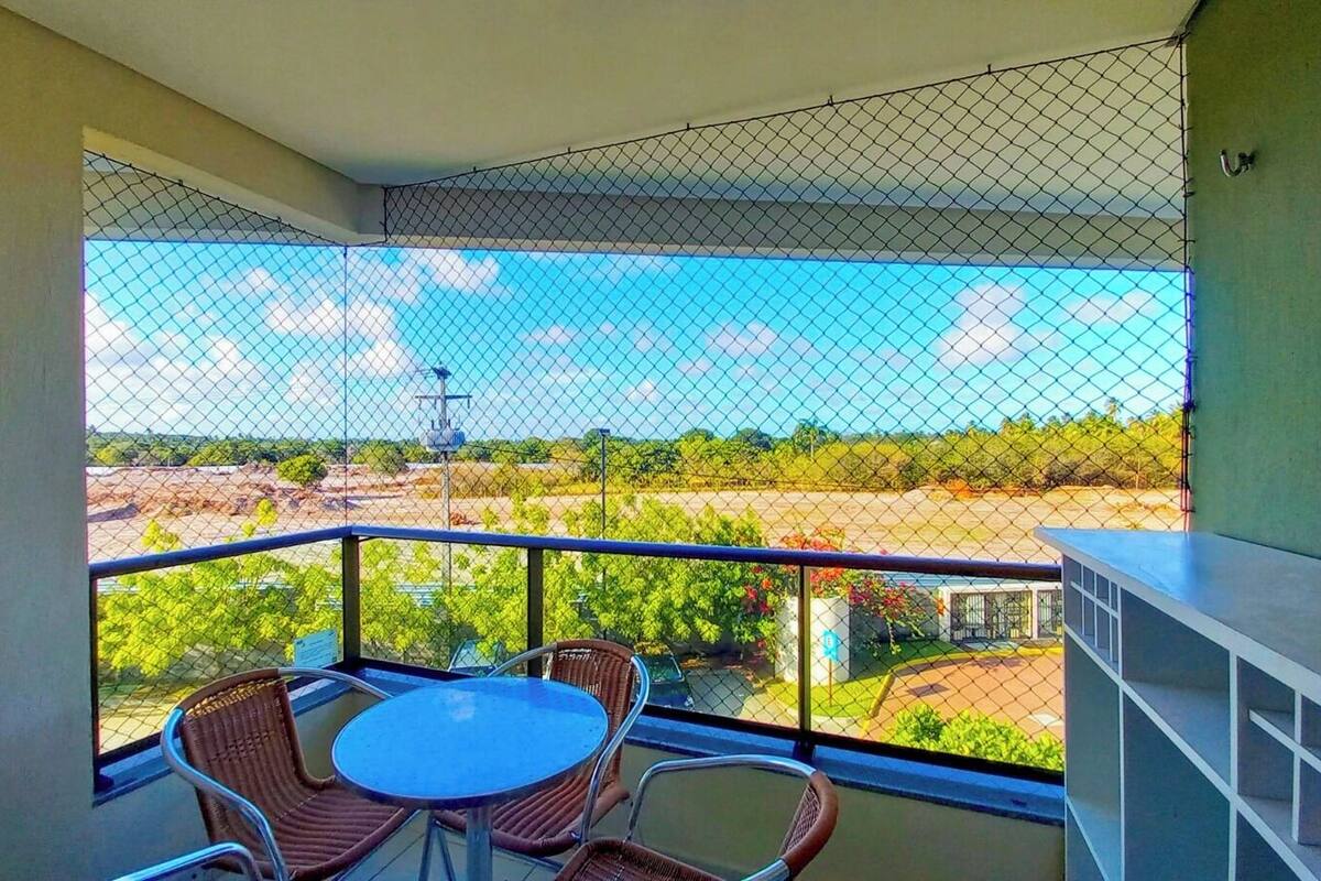 A balcony with a table and chairs, a view of a landscape with trees and a building, and a fence.