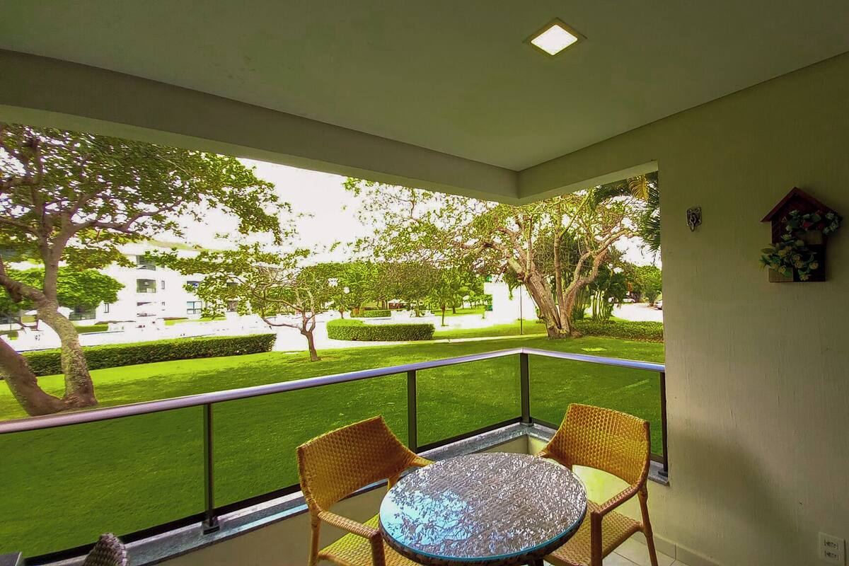 A balcony with two wicker chairs and a glass table, overlooking a well-maintained garden with trees and a hedge.