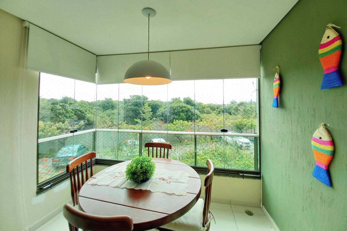 A dining area with a round table, chairs, a hanging light fixture, and a view through large windows.